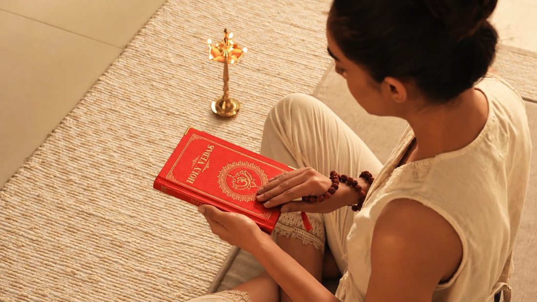 Frau auf einer geflochtenen Matte liest in einem roten Buch mit dem Titel Holy Vedas, daneben eine Messing-Öllampe, für Art of Vedas